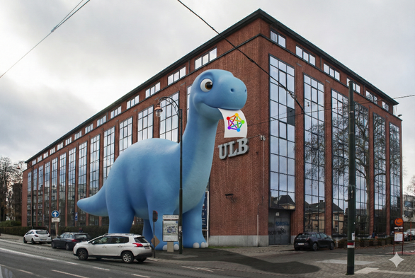 A photograph showing a giant, animated blue cartoon brachiosaurus standing next to a real-world brick university building with large glass windows and the letters “ULB” on the façade. The friendly-looking dinosaur holds a white paper with a colorful network diagram in its mouth. Parked cars and a street are in the foreground under an overcast sky.